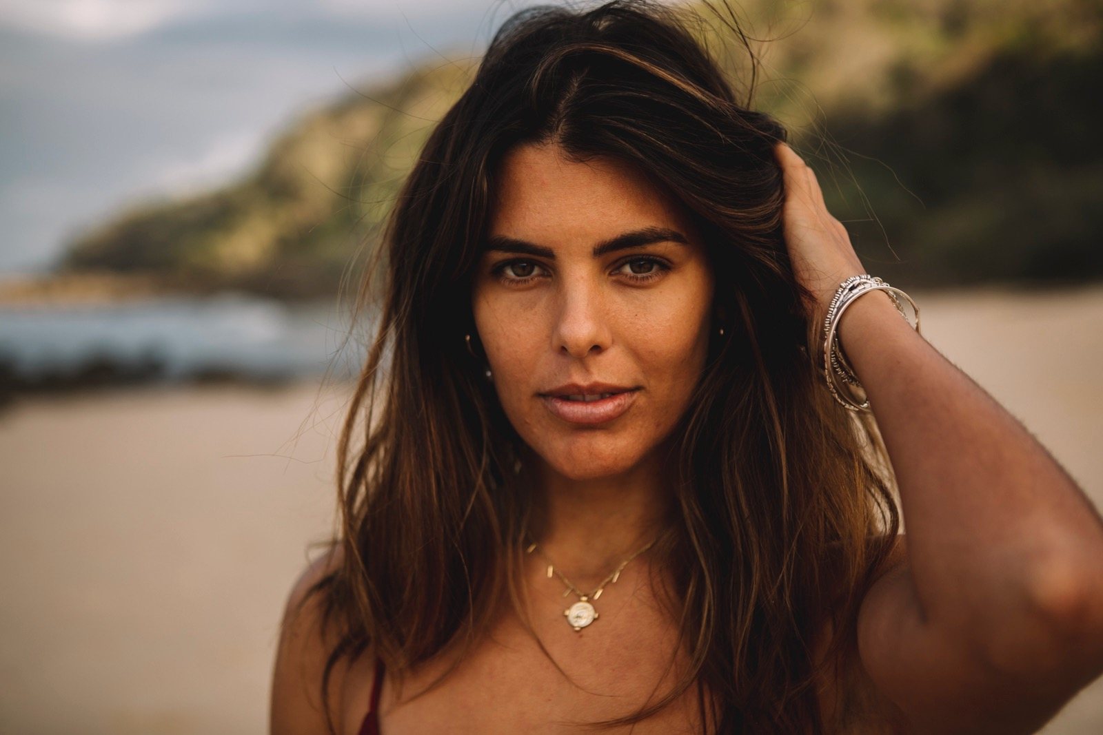 Portrait of Aquila Ana Bergstrom on a beach, one hand in her hair, a headland softly out of focus behind her