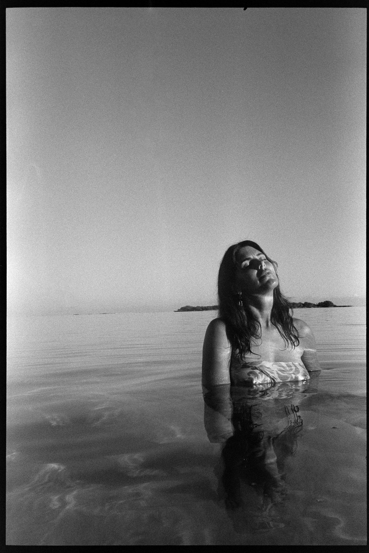 Black-and-white film photograph of Aquila standing in calm shallow water, head tilted back to the sun, a low island on the horizon behind her