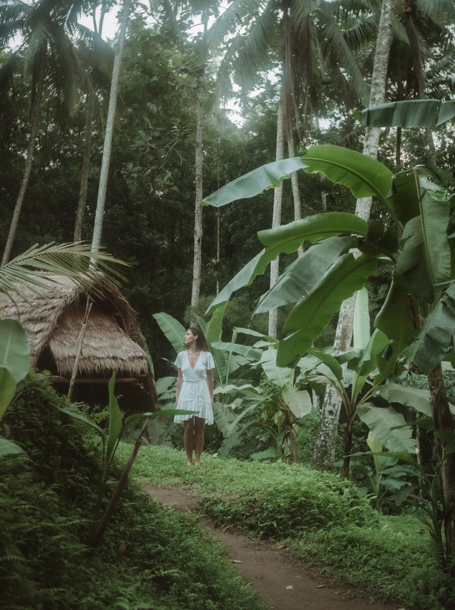Aquila in a white dress on a path through tall palms and banana leaves, a thatched hut beside her