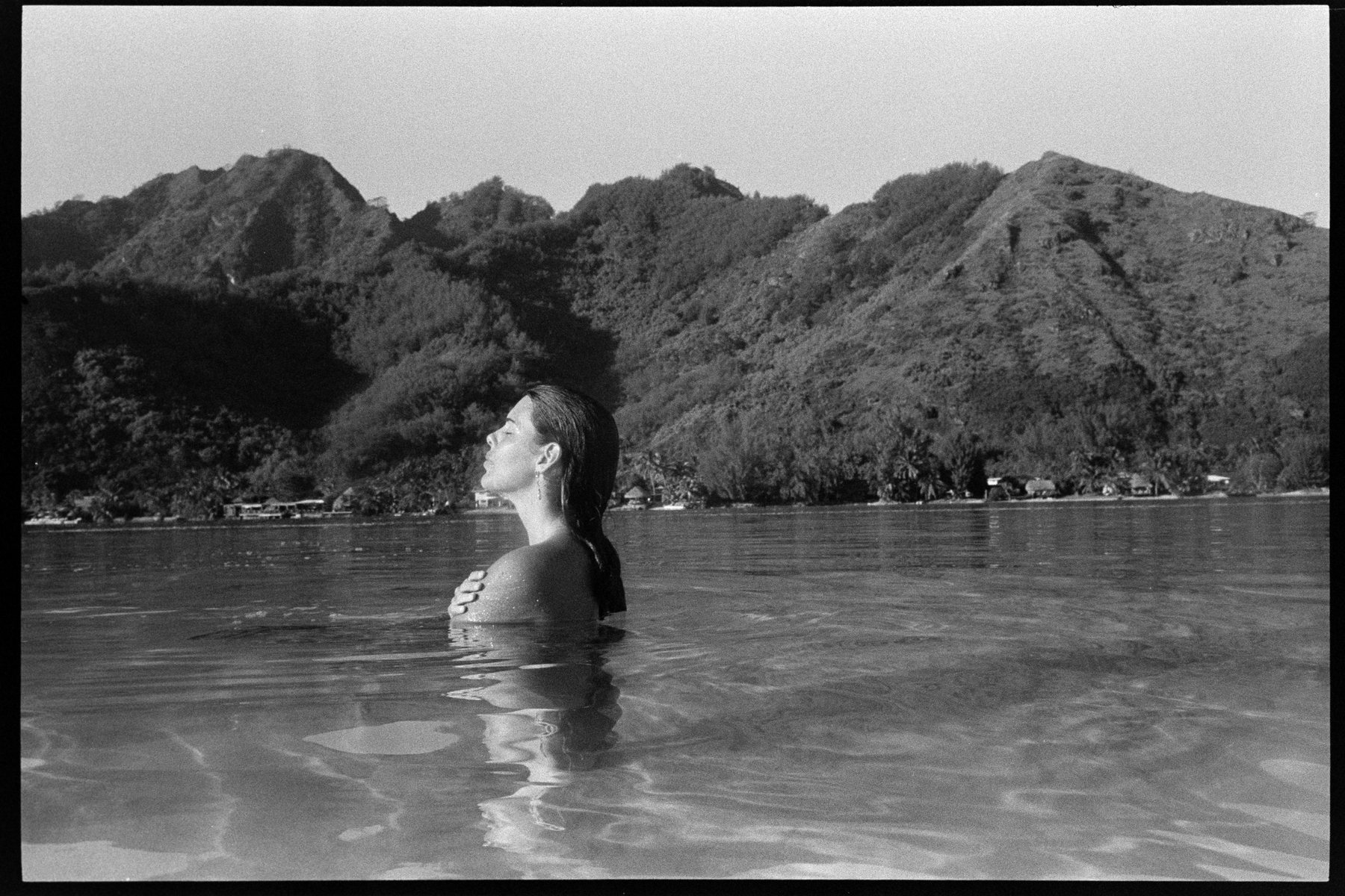 Black-and-white film photograph of Aquila standing shoulder-deep in a glassy lagoon, eyes closed and face to the sun, the steep green ridges of Moorea and a row of thatched bungalows behind her