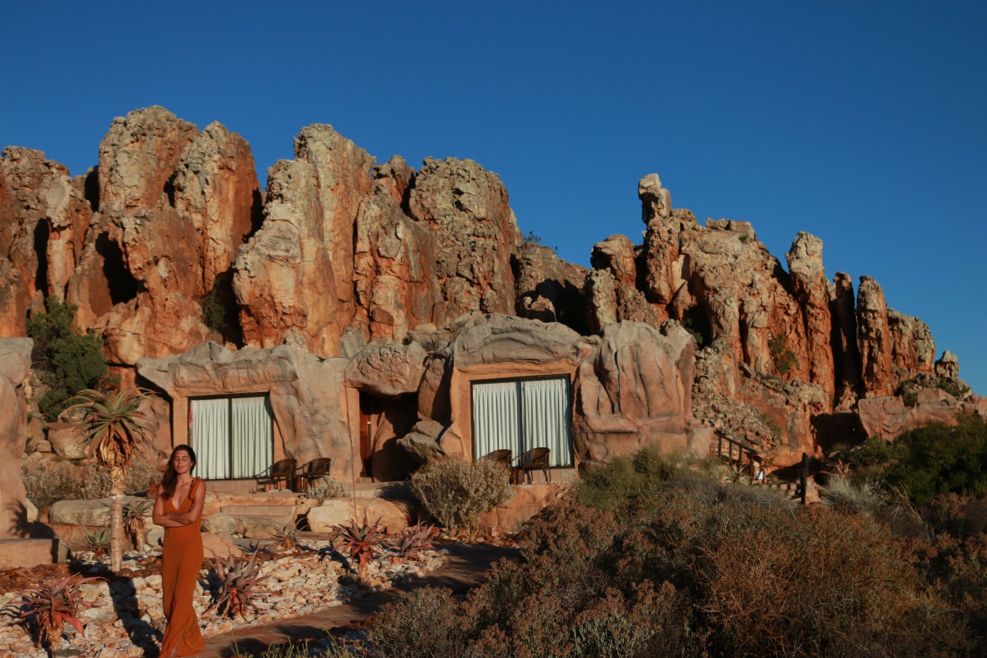 Aquila in a rust dress below the sandstone cliffs of Kagga Kamma at golden hour, cave suites built into the rock behind her